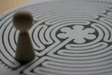A small wooden game piece standing at the edge of a printed circular labyrinth, its winding path curling toward a rosette at the centre.