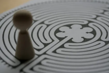 A small wooden game piece standing at the edge of a printed circular labyrinth, its winding path curling toward a rosette at the centre.