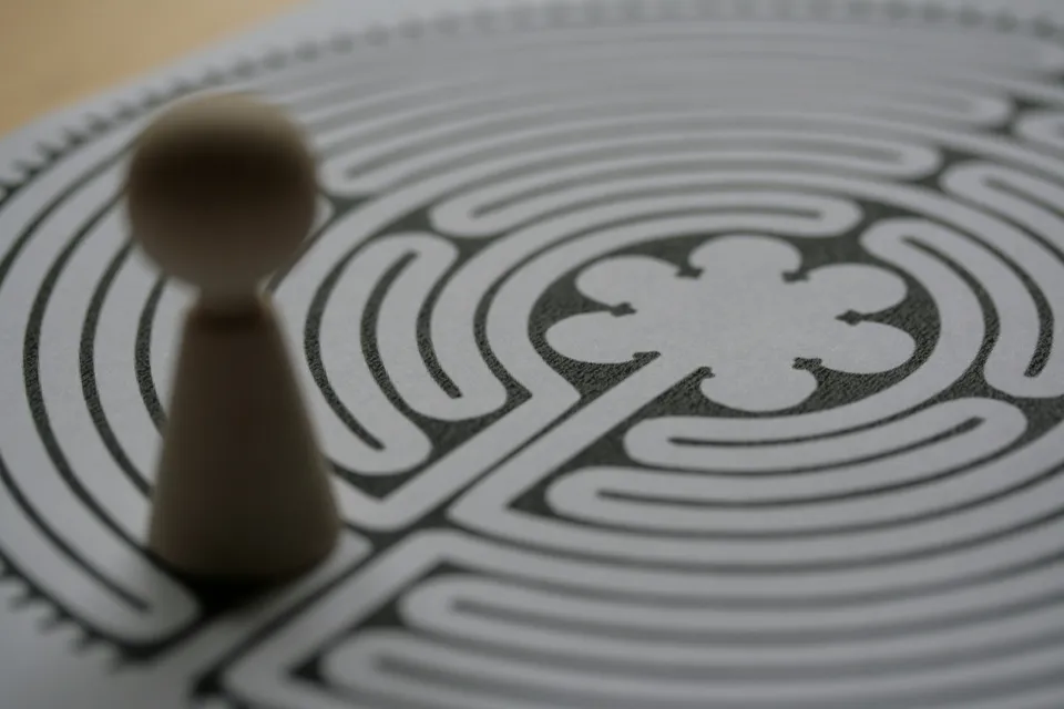 A small wooden game piece standing at the edge of a printed circular labyrinth, its winding path curling toward a rosette at the centre.